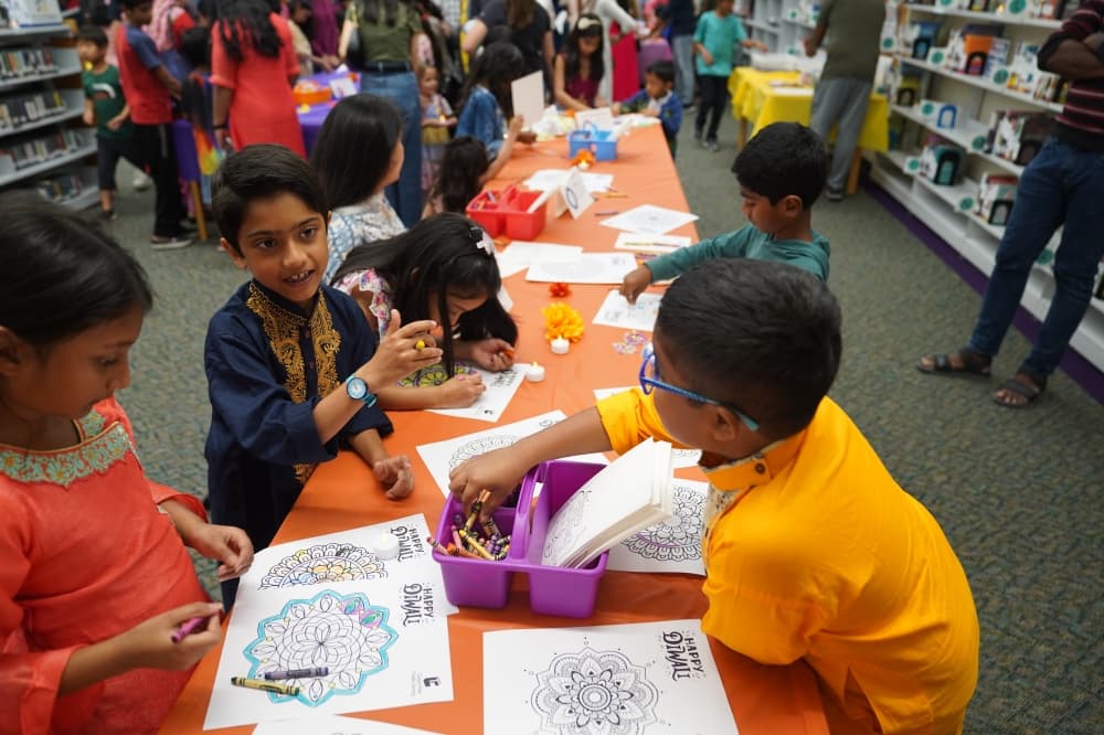 Children seated at a table with an orange tablecloth talk about their crafts.