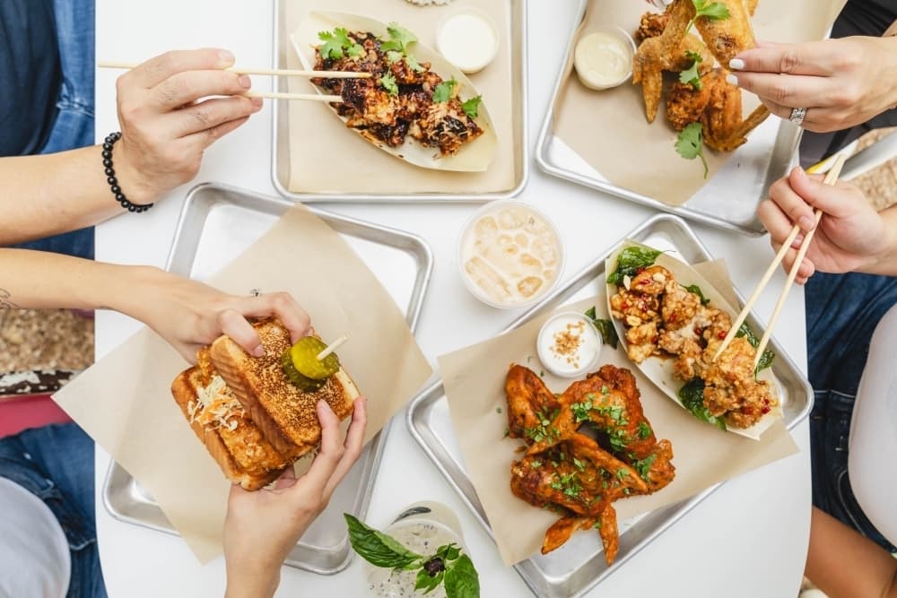 A white table shows the hands of four people eating four different dishes.