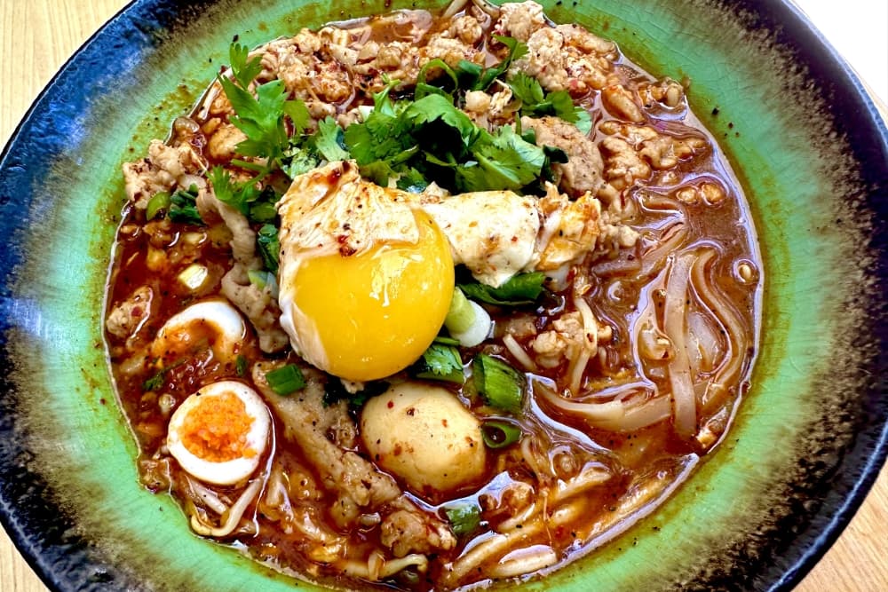A blue and green bowl holds a thick soup filled with noodles, vegetables and an over-easy egg.