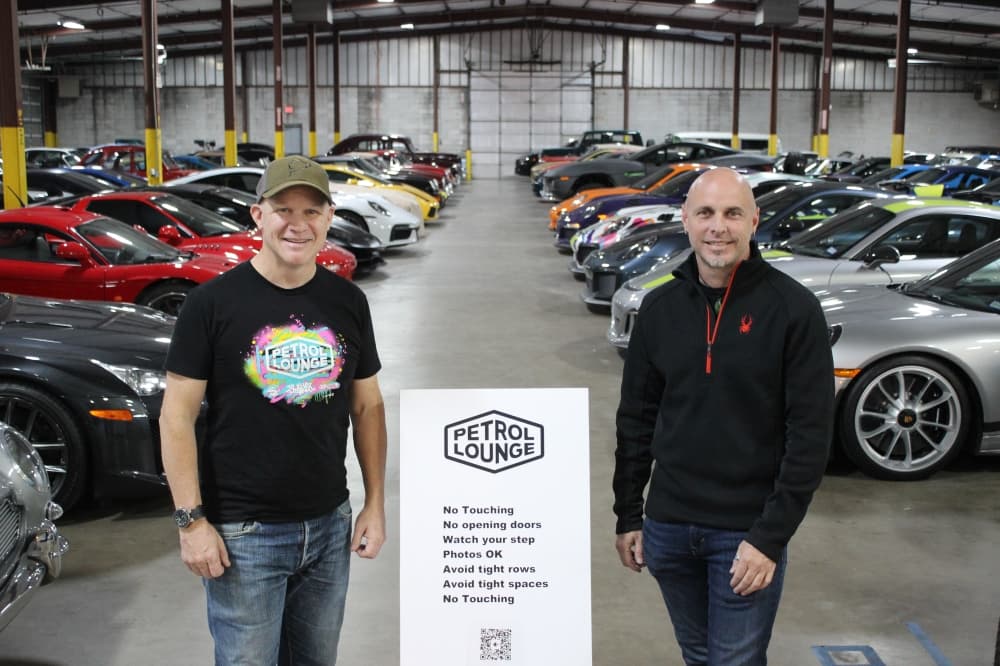 two men standing with "petrol lounge" sign in front of cars 