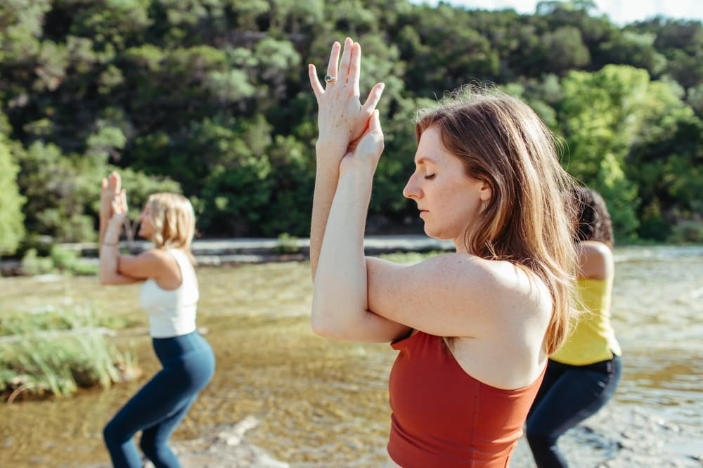 Three people practice yoga outside by a stream.