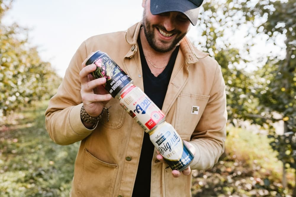 CEO Andrew Blake wears a cap and brown jacket and stocks a can of cider from each of the three companys while standing outside.