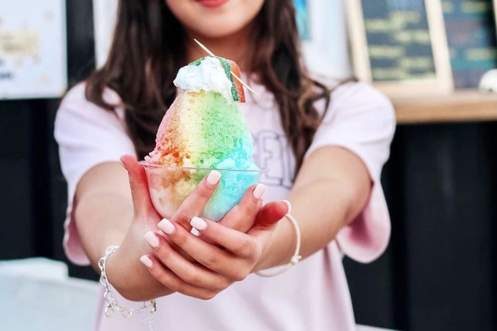 a girls hands holdout a rainbow colored snow cone with whip cream on top