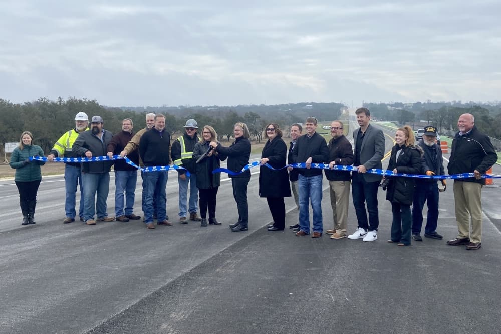 Several individuals stand in a line in front of a road to cut a blue ribbon.