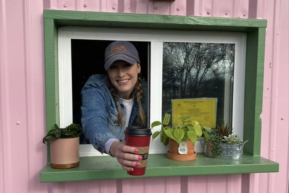 The owner hands a cup of coffee through the window while smiling.