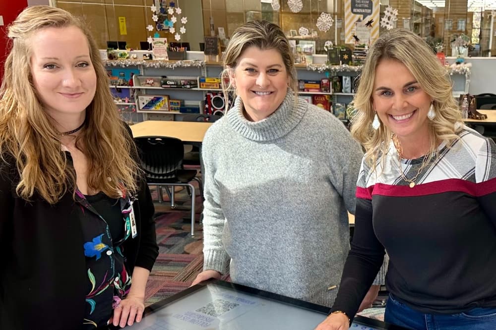 Three women post for a photo standing by a touchscreen digital table