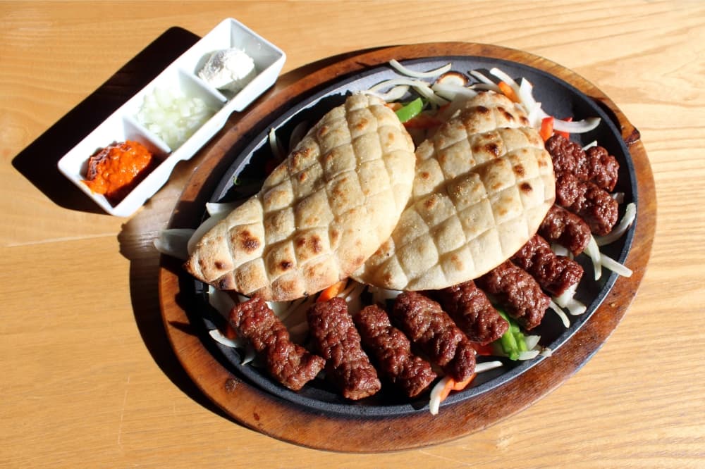Cast iron plate with beef sausages and two pieces of bread and a side of dipping sauces.