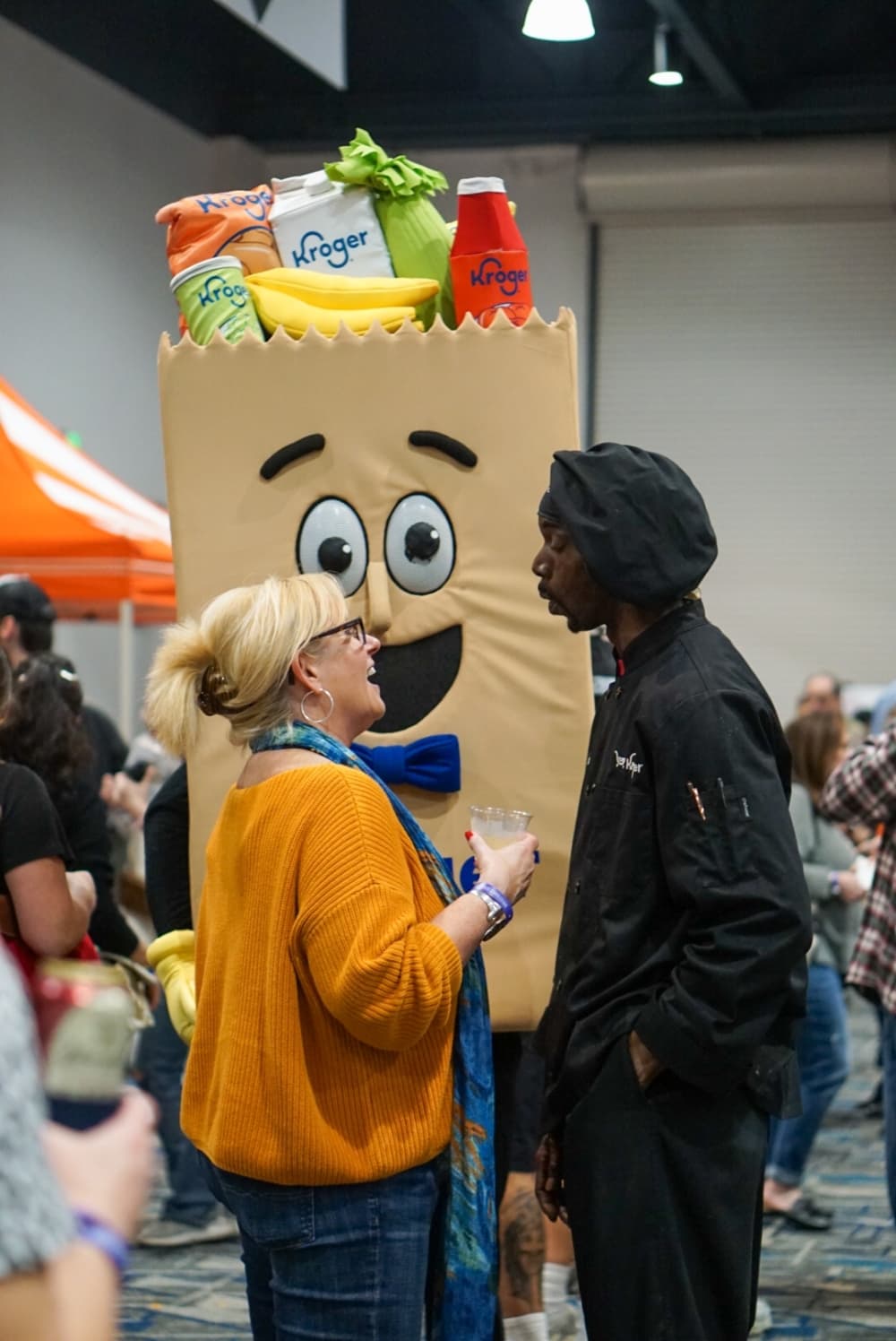A Kroger grocery bag mascot stands behind two talking people