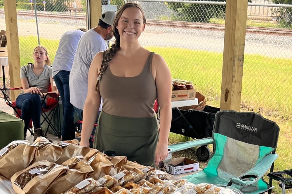 The owner smiles in front of a table of her products at a farmers market.