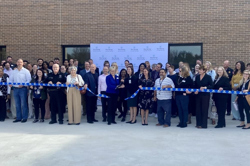 A group of people stand behind a blue ribbon while two people cut it with large scissors