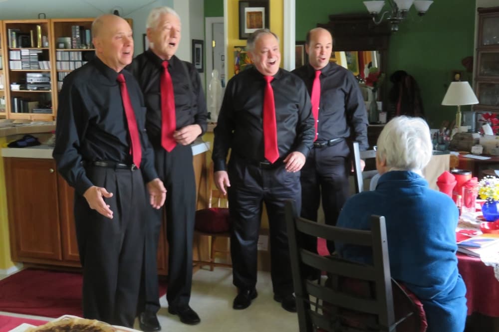 A barbershop quarter dressed in black with red ties sings to someone in their home.