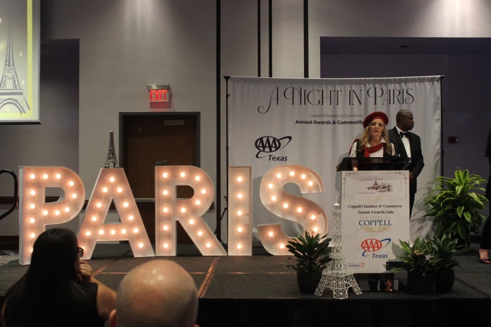 woman speaking at a podium in front of lit up signage that says paris