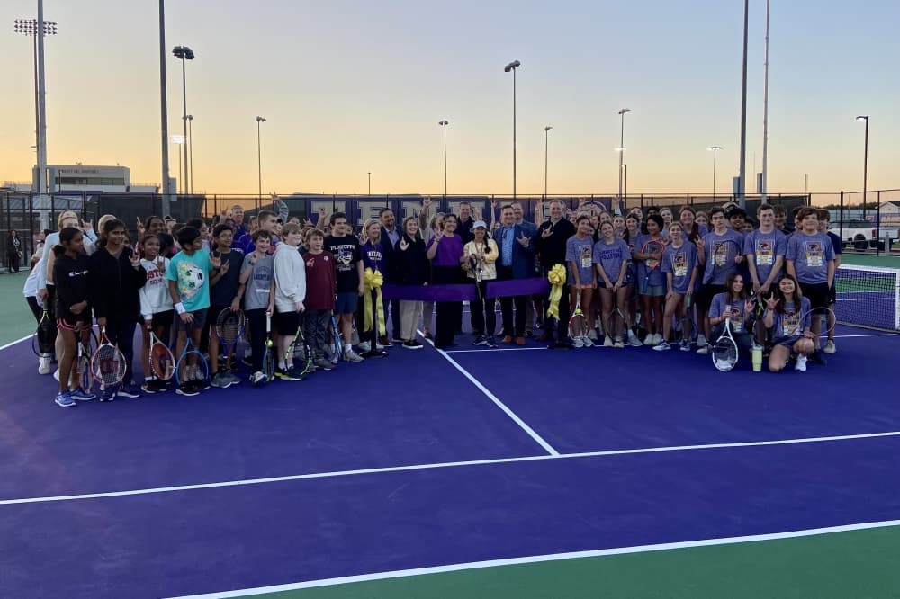 Dozens of students gather with tennis rackets on a purple tennis court while district officials cut a ribbon with big scissors.