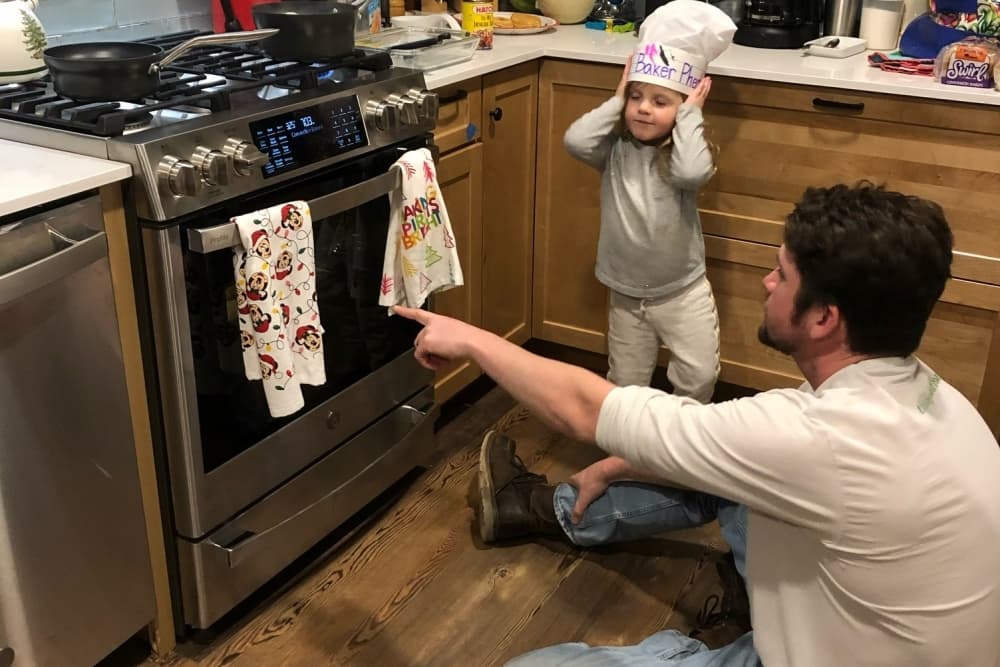 A small child wearing a chef's hat helps a man cook as he points at the oven in their kitchen.