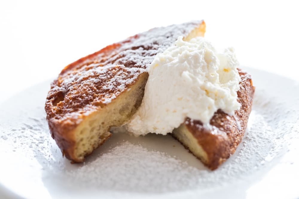 A white plate holds a deep-fried french toast with white whipped cream.