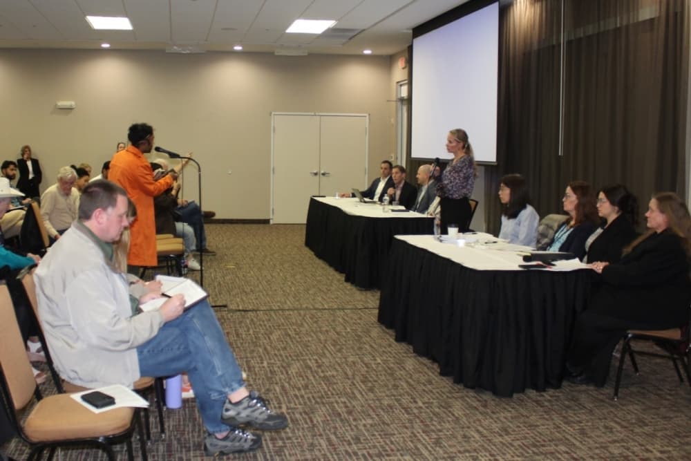 A speaker stands in front of a microphone while a panel of people are watching.