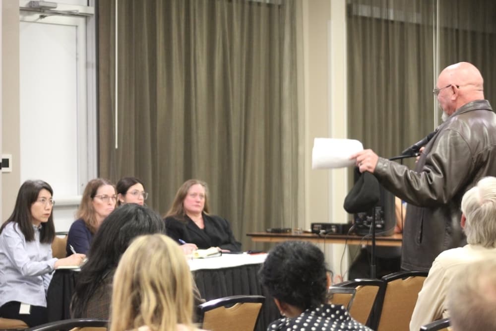A speaker stands in front of a microphone while a panel of people are watching.