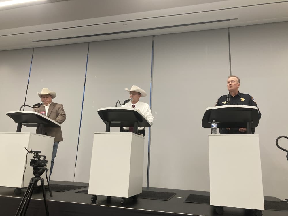 Three men stand at podiums; the two on the left are wearing cowboy hats