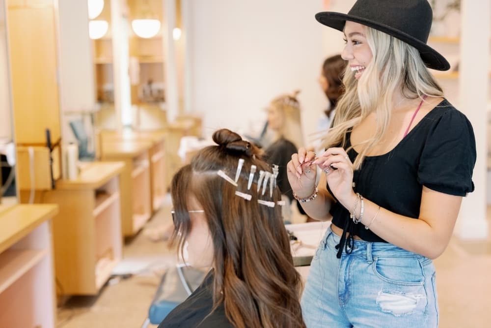 A woman in a black hat smiles in the mirror at another woman sitting down receiving a hair service.