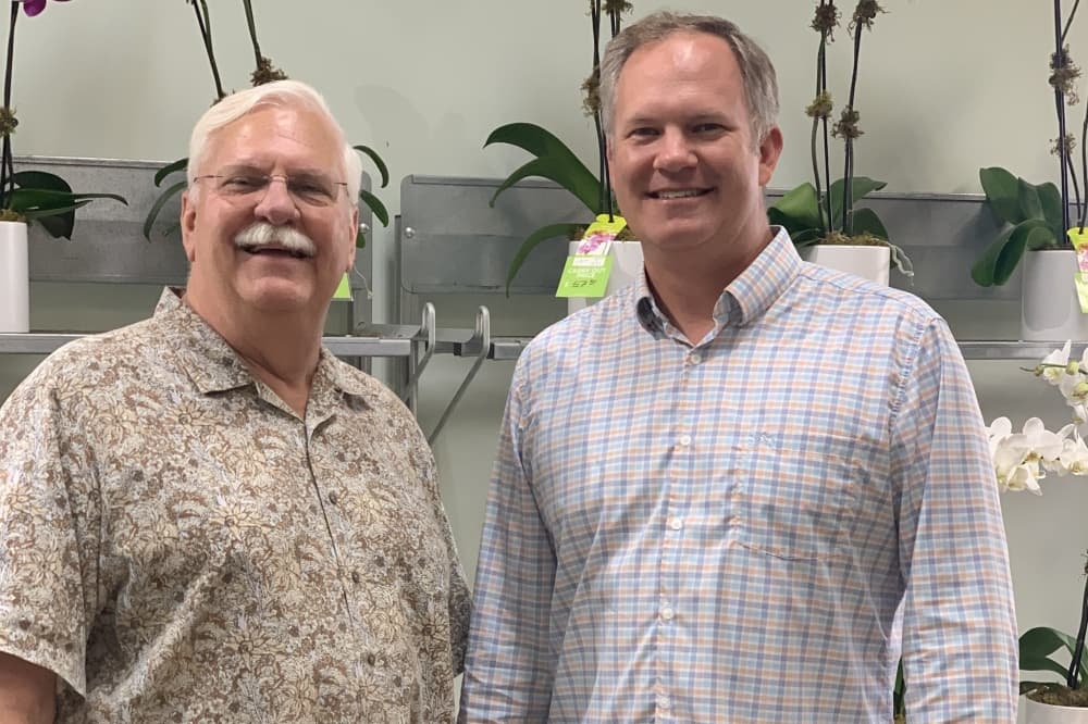 Two men stand side-by-side smiling in front of the flowers at the shop.
