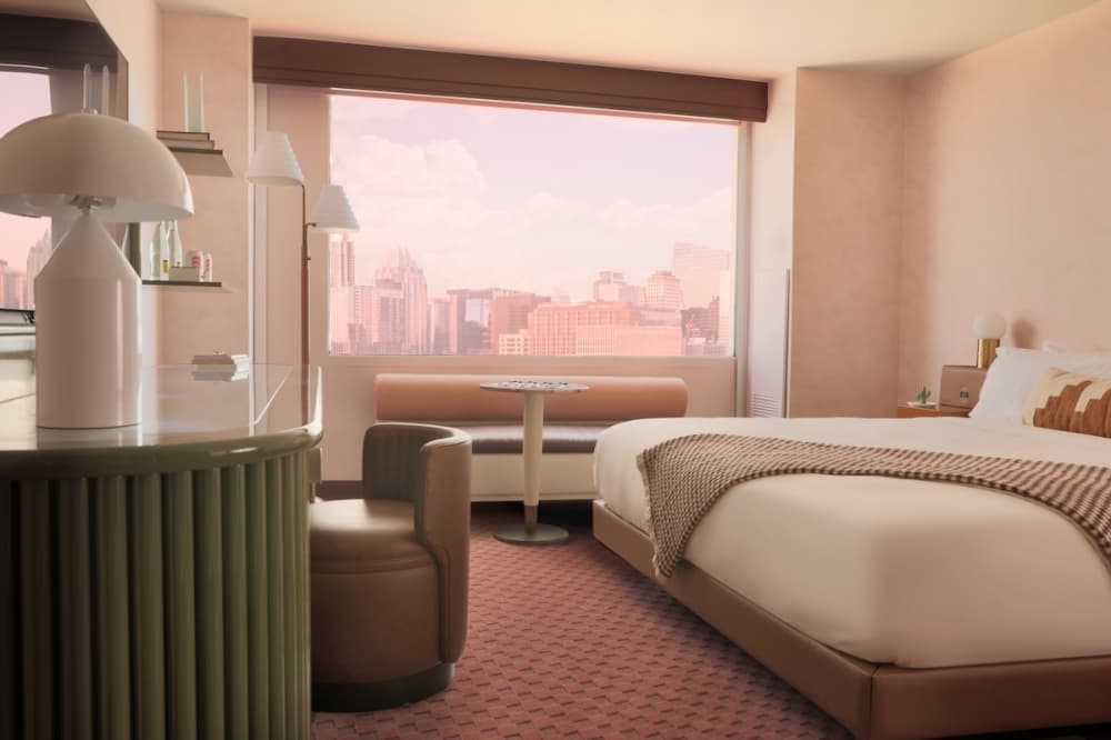 A guest room is shown, with a look at the bed and views of the city in the background through one of the windows.