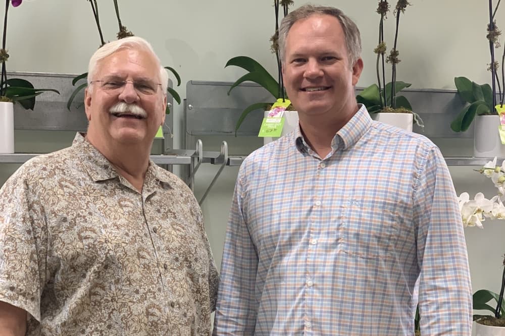 The two men pose side-by-side in front of a wall with plants for sale.