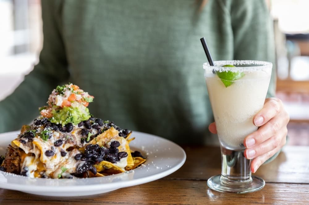 A big plate of nachos with many toppings is pictured next to a frozen margarita.