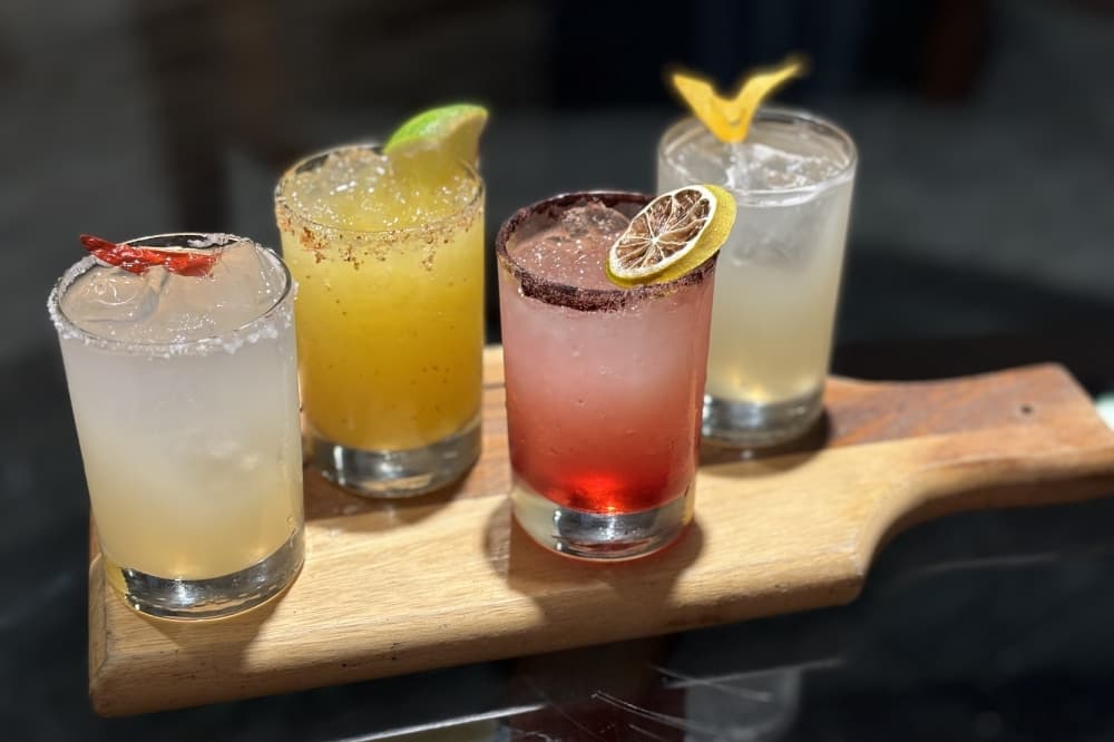Four margaritas of varying colors are pictured on a wooden flight paddle. 