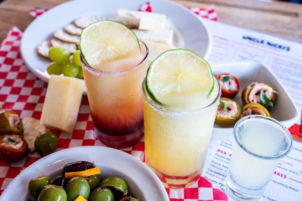 A red and a yellow margarita are shown on a table surrounded by food, next to a shot glass.