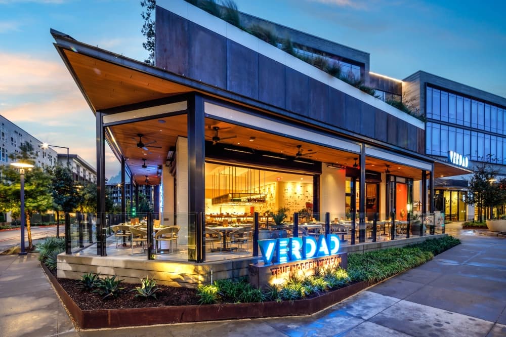 An exterior shot of the restaurant is shown featuring a look at the outdoor patio areas and a blue neon sign that reads "Verdad."