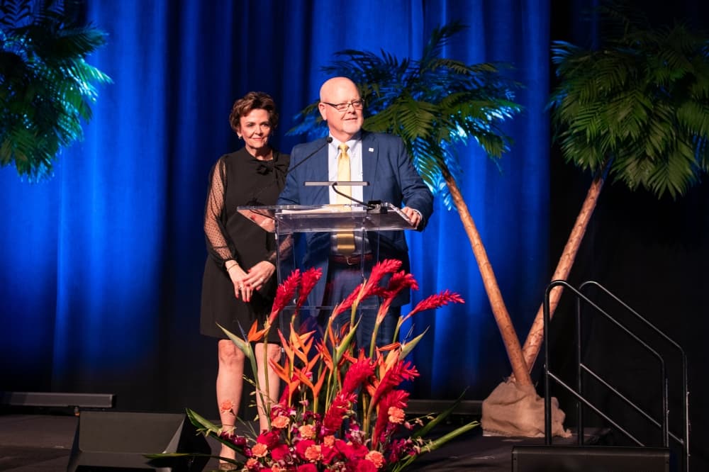 A man and a woman speak at a podium flanked by artificial palm trees.