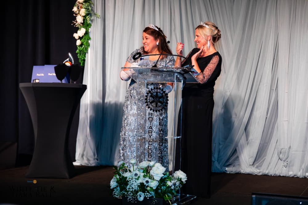 Two women stand at a podium wearing tiaras.