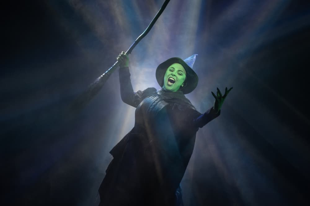 The green witch holds her broom and poses in front of a bright light.