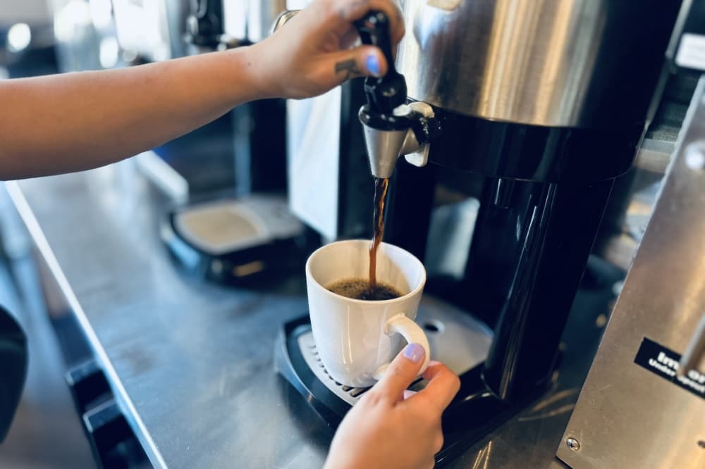 The barista's hands are shown pouring a cup of black coffee from the machine.