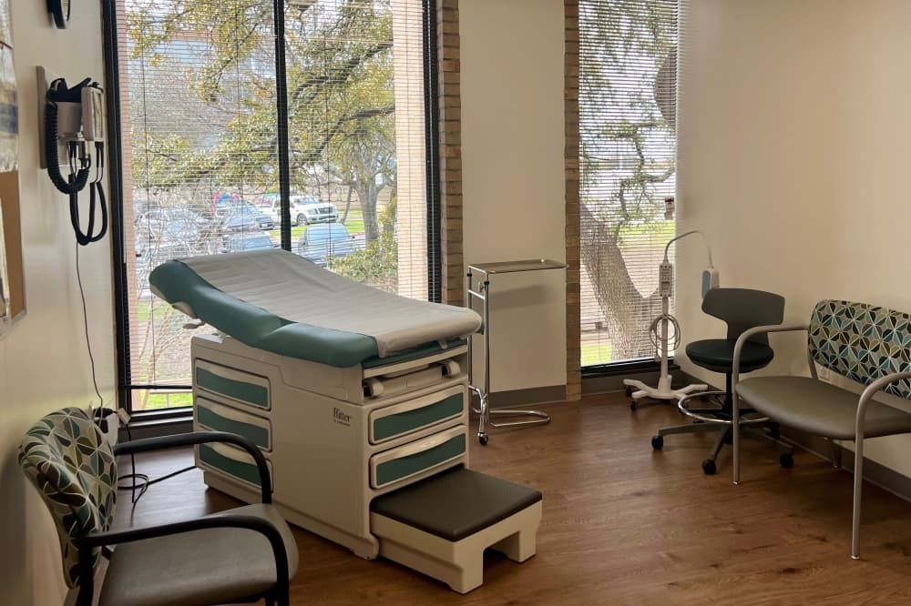 An empty room with a medical exam table and chairs with windows showing the trees outside in the background.