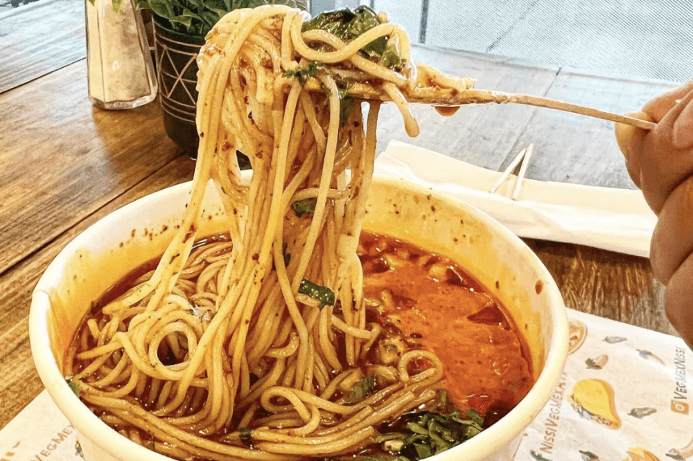 A person holds a fork above the ramen birria with noodles stringing down.