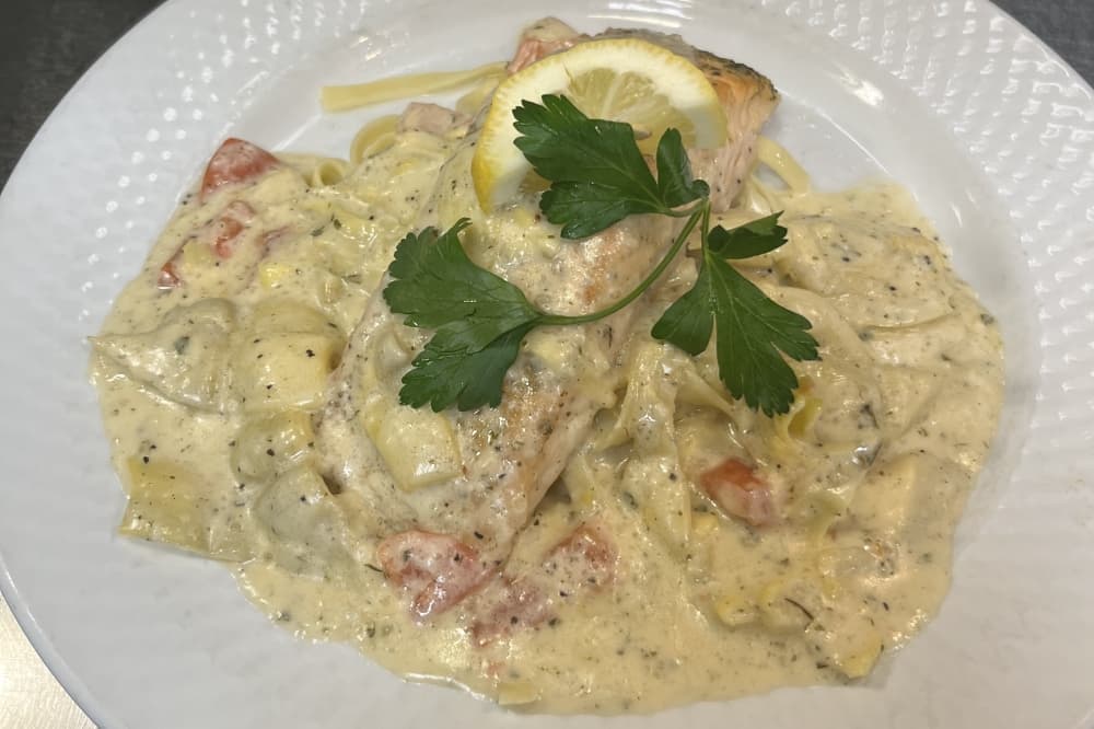 A salmon filet is covered in a cream sauce and garnished with parsley and a lemon slice.