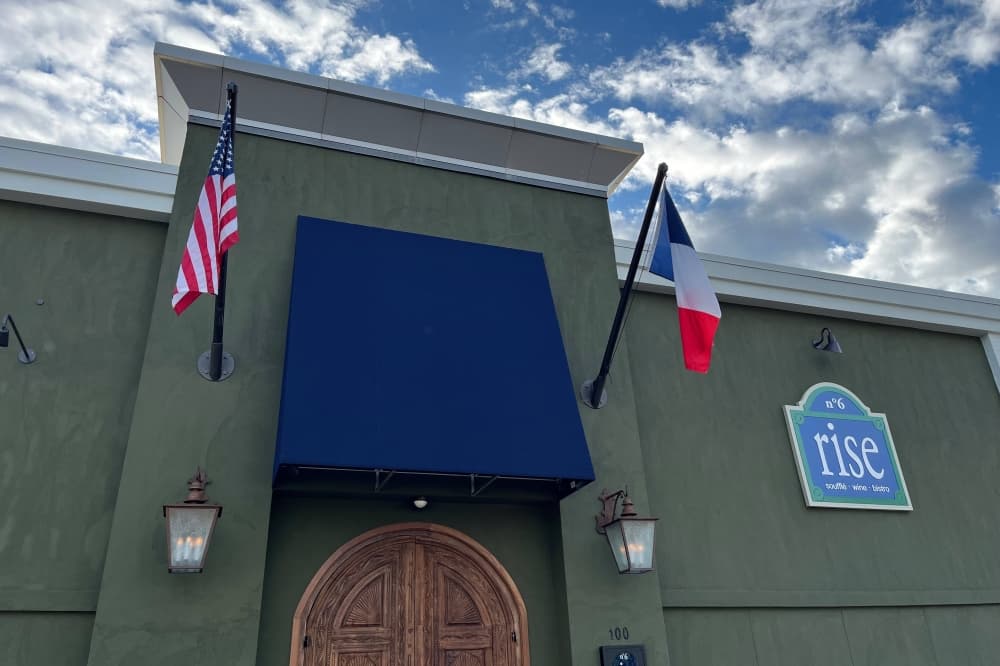 Front of a building with Rise logo and American flag and French flag flanking blue awning over brown door. 
