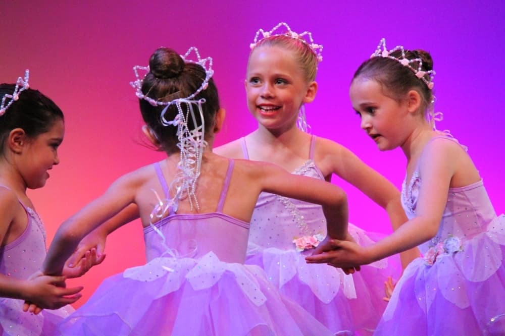 Four children are shown holding hands on stage dressed in ballerina costumes