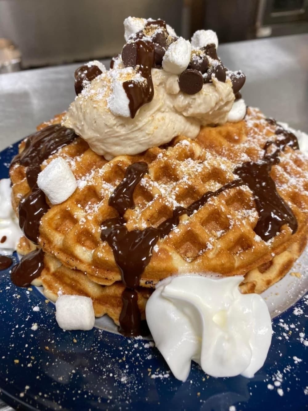 A Belgian waffle with whipped cream, chocolate sauce, powdered sugar and marshmallows is shown.