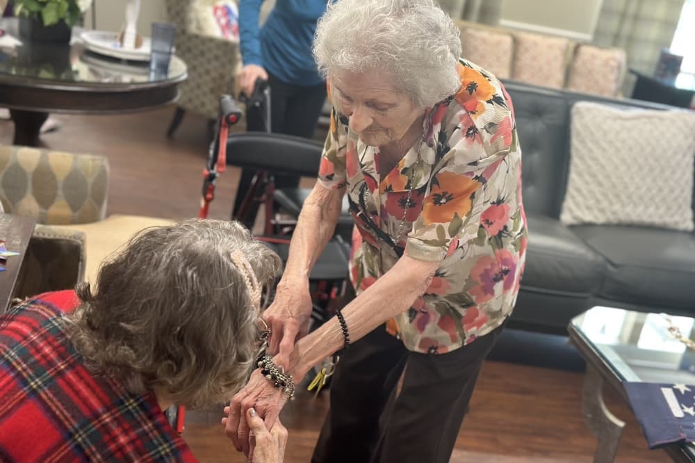 A woman leans down to show another woman, who is seated, her bracelet.