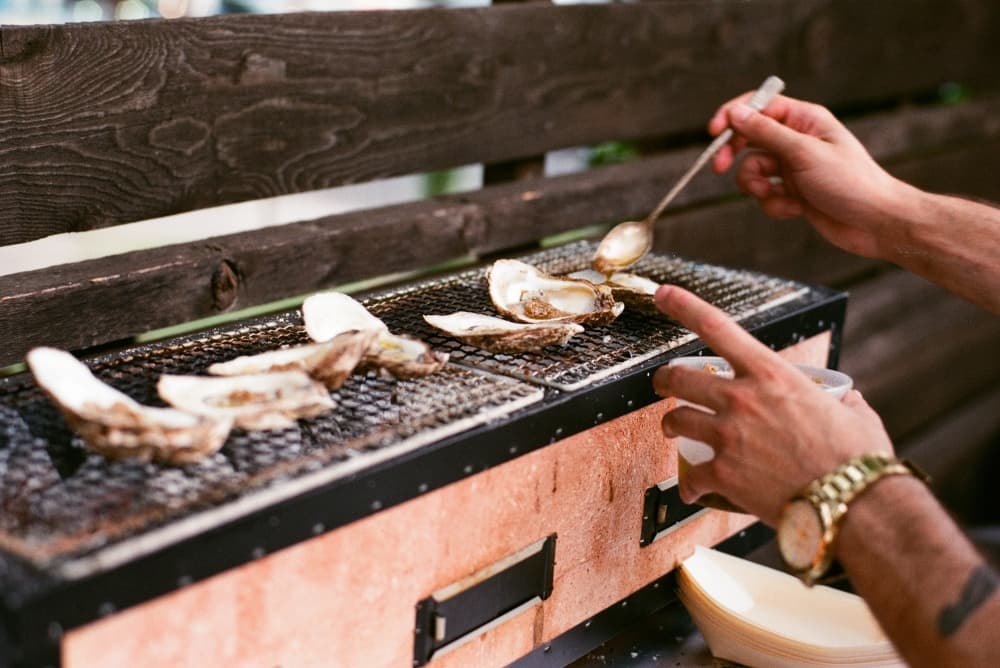 A grill is lined with oysters while someone bastes them.