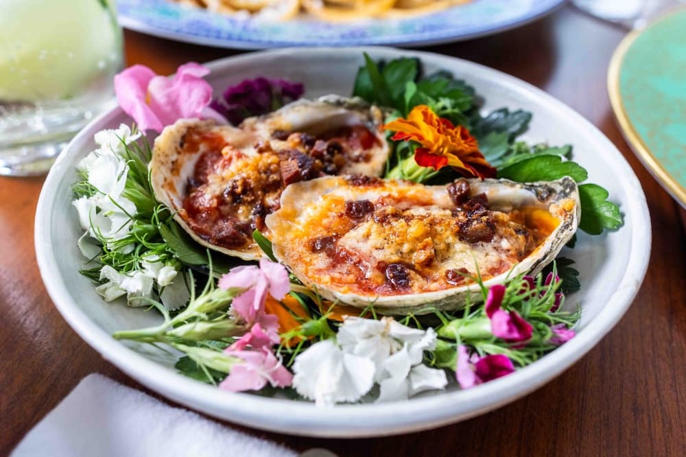 A white bowl holds an oyster dish with purple and white edible flowers.
