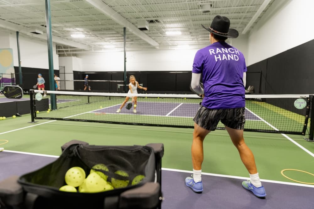 Two people play pickleball—in the foreground, a person wearing a black cowboy hat and a purple shirt reading "Ranch Hand" is shown; in the background, a person in white sportswear is shown.