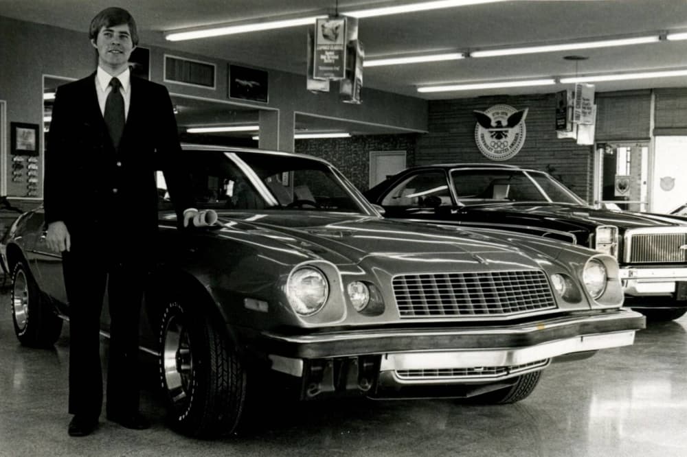 Black and white photo of a man standing next to a car in a car dealership showroom.