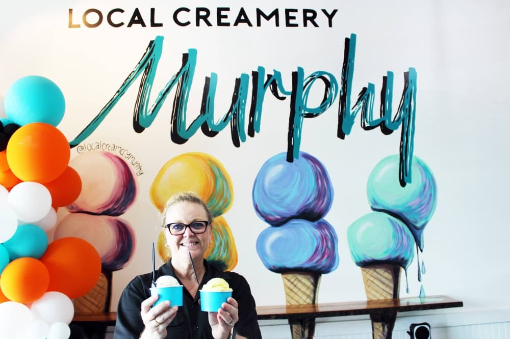 Female holding two cups of ice cream in front of wall with Local Creamery Murphy signage. 
