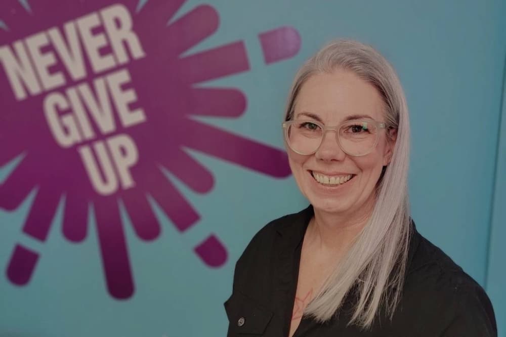 Female standing in front of a wall with 'Never Give Up' signage on a wall behind her. 