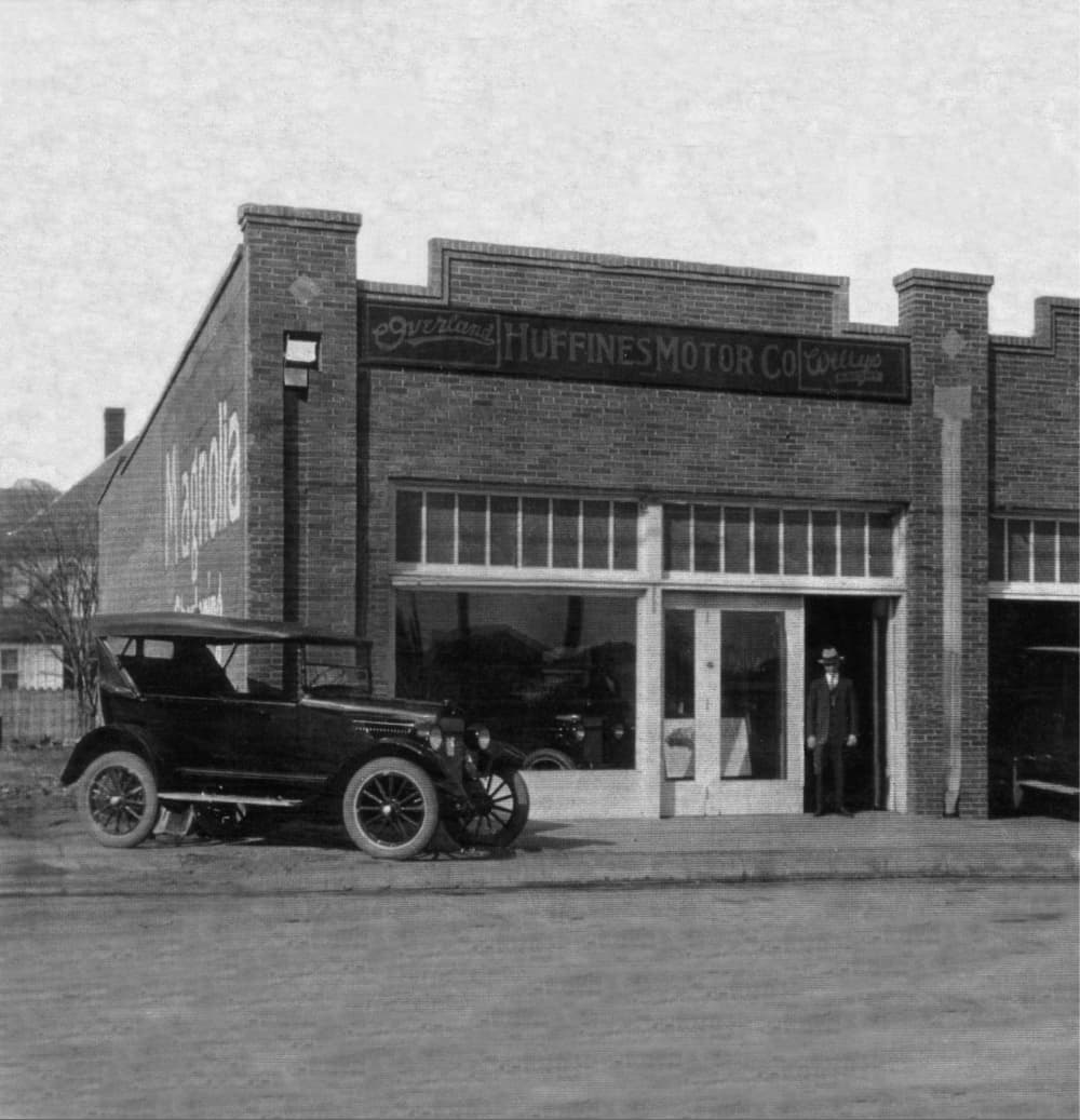 Black and white photo of a old building with Huffines signage and antique car parked in front of the building. 