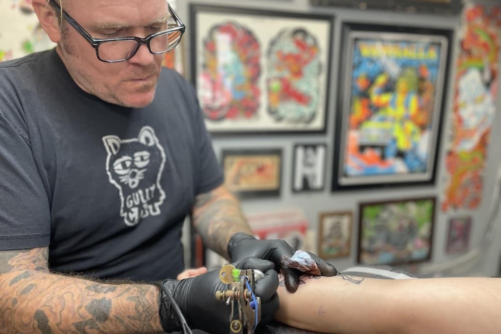 Image shows Joel Valkner tattooing a client.