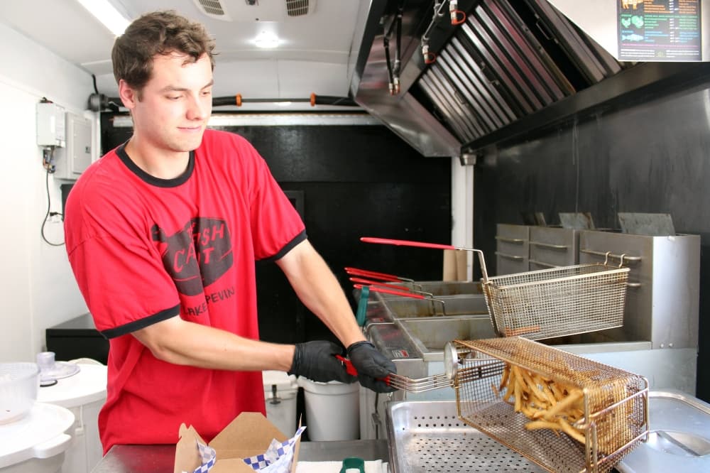 Male wearing The Catfish Hut red shirt while dumping basket of fries on a metal tray. 